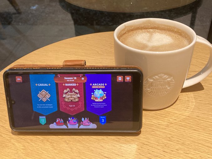 A smartphone displaying a Season 18 game menu with Casual, Ranked, and Arcade game modes sits next to a Starbucks coffee cup on a wooden table.