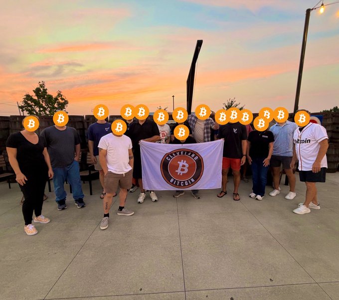 A group of people stand outdoors at sunset holding a "Cleveland Bitcoin" flag, with Bitcoin logo emojis covering their faces.