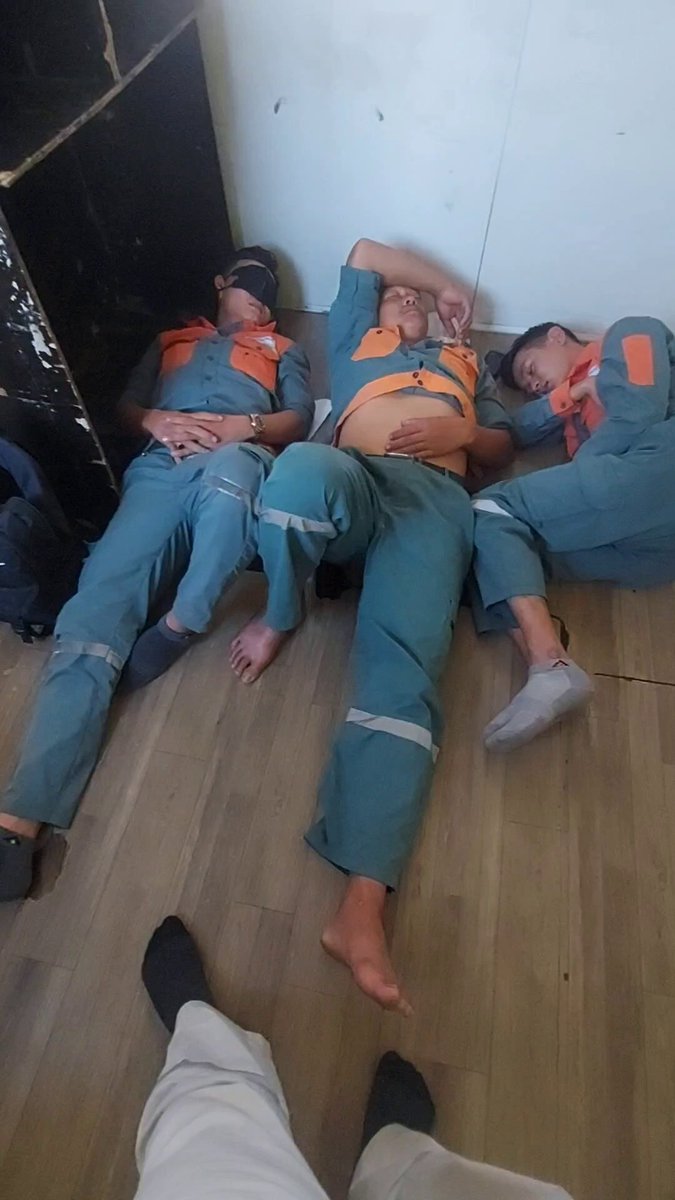 The image shows three people lying down on the floor of an industrial setting with their legs crossed. They appear to be taking a nap or resting, possibly after working long hours. The background features a wall that has been stripped of its paint, giving the room a worn and aged appearance. A person is standing in front of this stripped wall, observing the scene.