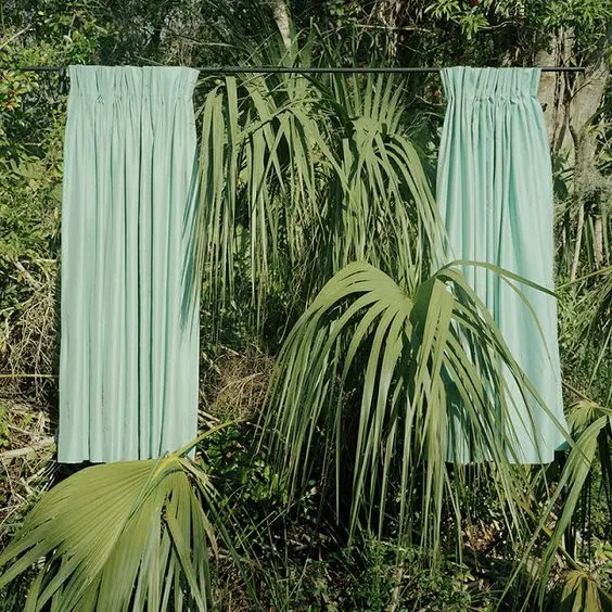 Two pale mint-green pleated curtains hang from a rod outdoors, framed by green palm-like foliage in a tropical setting.