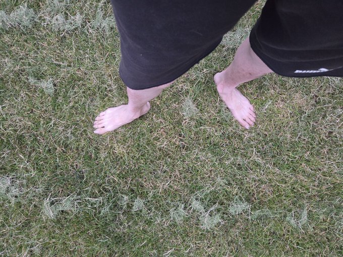A person wearing black shorts stands barefoot on a patch of grass mixed with dry clippings.