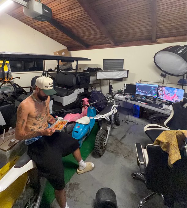 A shirtless tattooed man in a cap sitting in a garage eating from a takeout container, surrounded by a golf cart, motorcycles, and a gaming setup with multiple monitors.