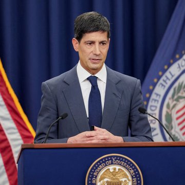 A man in a dark suit and tie stands at a podium with the seal of the United States Federal Reserve, speaking into microphones with an American flag in the background, illustrating the breaking news about Fed officials warning that more rate hikes may be needed if inflation doesn't decline—a bearish signal for risk assets.
