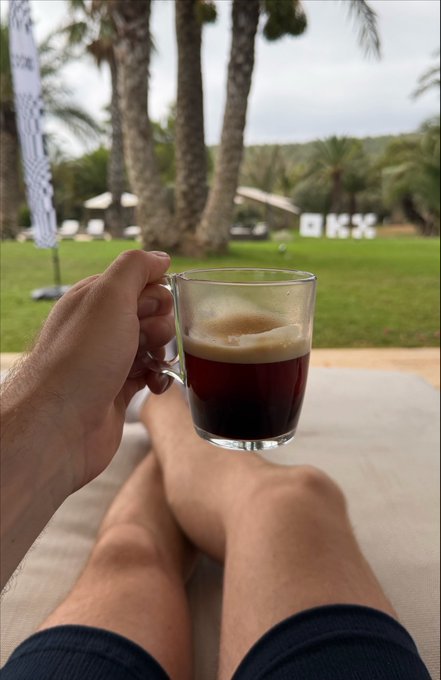 A person relaxes on a lounge chair at the OKX Creator Retreat in Ibiza, holding up a glass of espresso toward palm trees and "OKX" signage in the background, perfectly capturing the laid-back luxury of day 2.