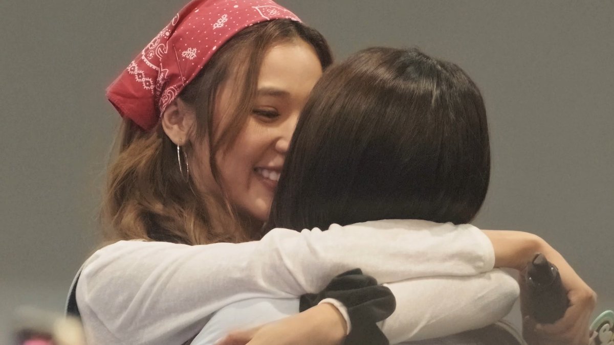 A woman wearing a red bandana smiles joyfully while hugging another person with a dark bob haircut.
