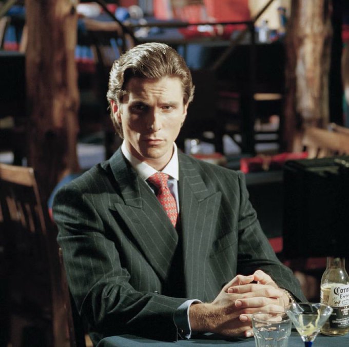A man in a dark pinstripe suit with a red patterned tie sits at a table with a martini glass, looking directly at the camera with a serious expression, featuring a Corona Extra bottle on the table.