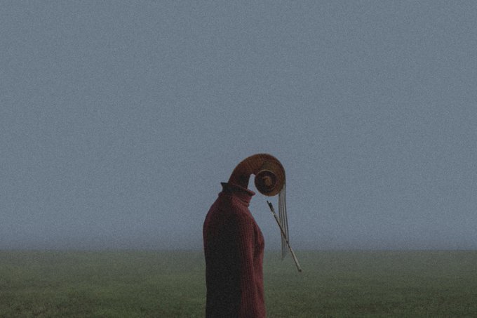 A grainy, atmospheric photo of a hooded figure in a red cloak standing alone in a foggy field, wearing a curious hat with hanging strings.