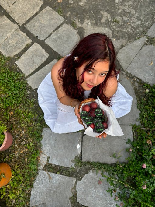 A young woman with dark curly hair stands on a stone pathway holding a basket of plums, looking up at the camera from a high angle.