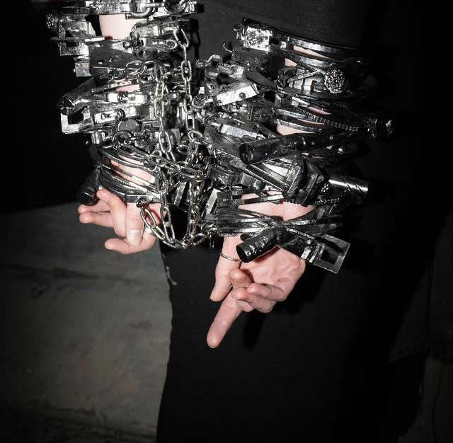 A person's wrists and hands are bound with an elaborate, heavy assemblage of black-painted chains, metal cuffs, and locks, creating a striking avant-garde bondage-inspired aesthetic against a dark background.