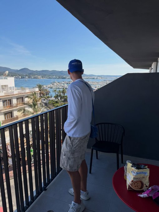A person stands on a balcony overlooking a marina and ocean view, wearing a blue cap, white long-sleeve shirt, and white shorts, with a bag of 'Arnott's Kingstons' biscuits on a red table in the foreground.