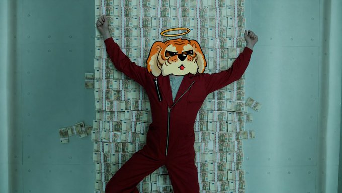 A person in a red jumpsuit lies on a pile of money with a cartoonish dog mask featuring an orange halo and sunglasses covering their face.