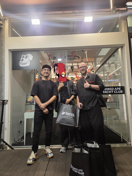 Three men pose in front of a 'Bored Ape Yacht Club' storefront, holding black BAYC shopping bags.