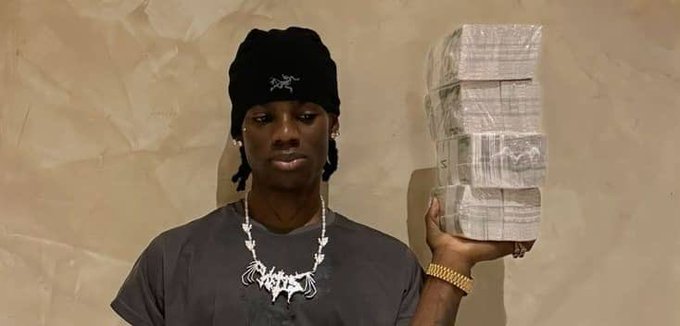 A person wearing a black beanie and a dark shirt with a chain necklace holds up a tall stack of paper currency, looking toward the camera.