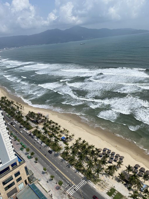 An aerial view of a tropical coastline featuring a sandy beach lined with palm trees, a road with parked cars, crashing ocean waves, and mountains visible in the background under a partly cloudy sky.