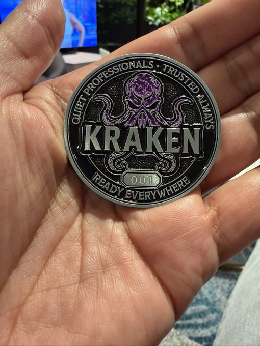 A hand holds a metallic "Kraken" challenge coin featuring a purple octopus design and the text "Quiet Professionals • Trusted Always • Ready Everywhere" with the number "001," likely representing the popular cryptocurrency exchange Kraken rather than relating to the QWO ticker.