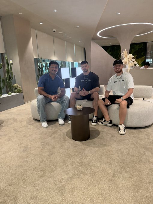 Three young men smile while seated on a white sofa in a modern lobby with a curved coffee table and cactus decor in the background, posing for a casual photo likely after meeting, fitting the laid-back vibe of the "GNiggabull" post.