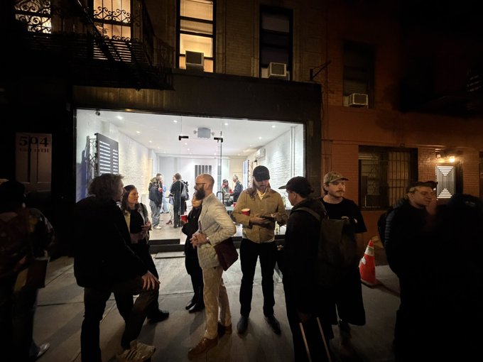 Nighttime photograph of a crowd of people gathered outside an art gallery or event space at 604 Main Street, with individuals holding red cups and engaging in conversation on the sidewalk.