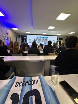 A photo taken from the audience perspective at an event, showing a person holding a 'DEEPCOIN' jersey with the number 20 in the foreground, while other attendees watch a presentation on screens at the front of the room.