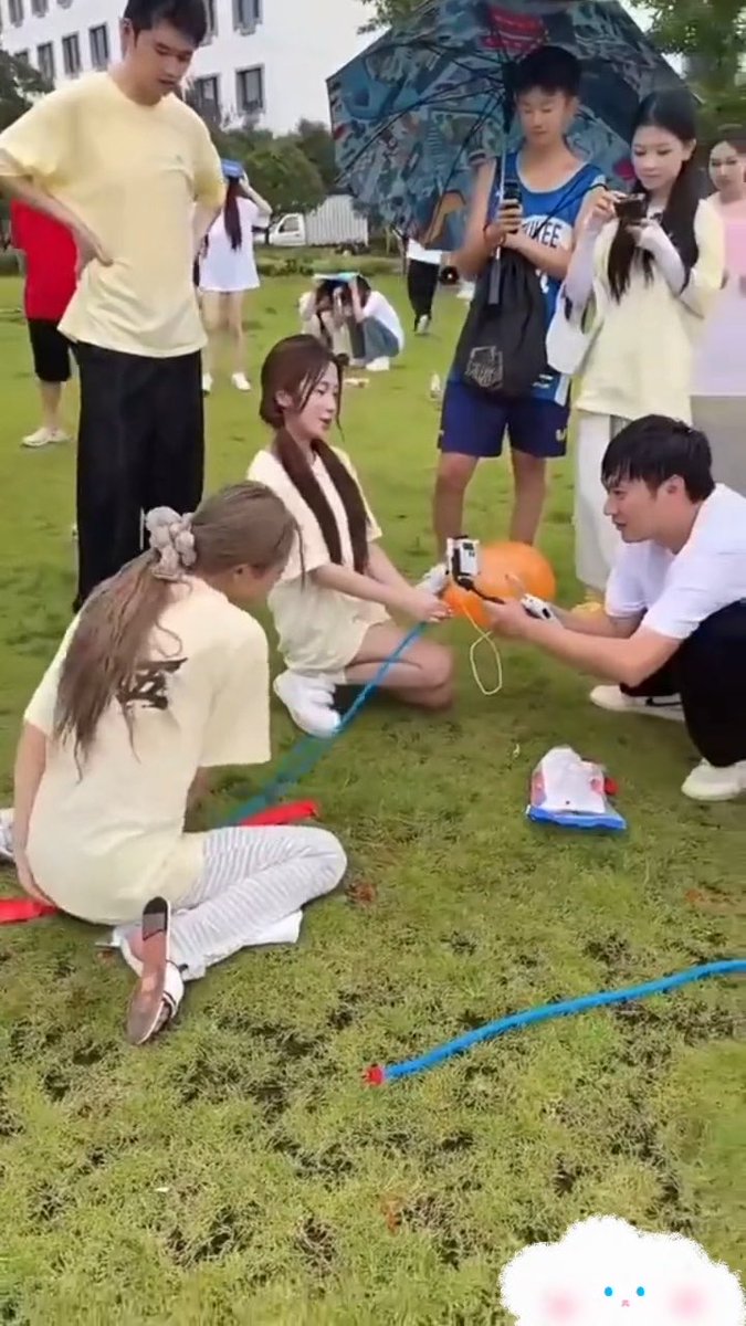 A group of young people gathered on a grassy field, with a young man kneeling to hold a pump connected to an orange balloon while others watch and record with phones.