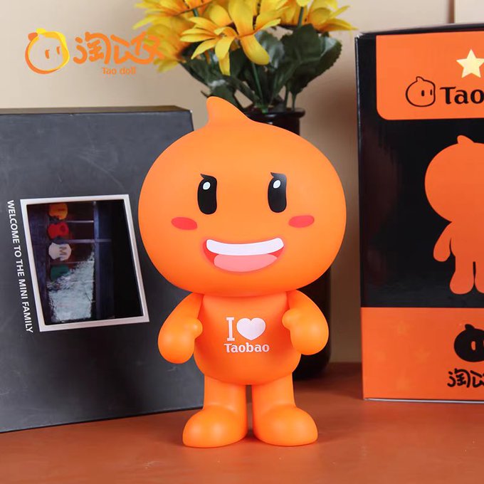 Orange Tao Doll figurine (淘公仔) wearing an 'I love Taobao' t-shirt, displayed next to its branded packaging box on a wooden surface with yellow flowers in the background.