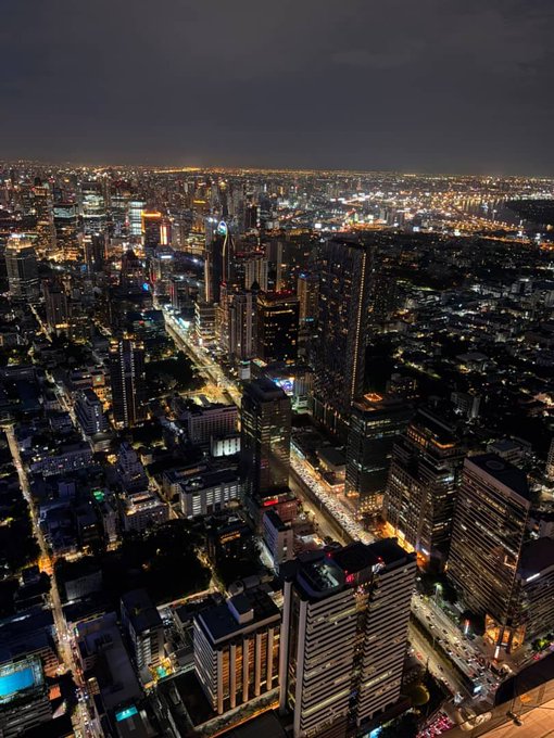 An aerial nighttime view of a sprawling, brightly lit city skyline with numerous illuminated skyscrapers and busy streets stretching to the horizon.