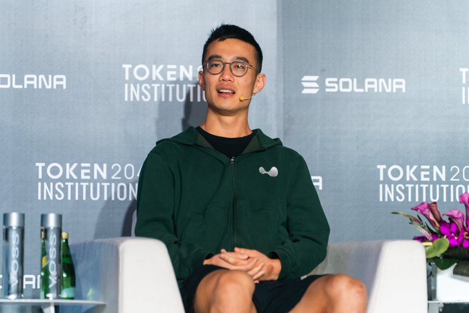 A young man in a green hoodie and glasses speaks into a headset microphone while seated on stage, with the "TOKEN2049 INSTITUTIONAL" and "SOLANA" branding visible on the backdrop behind him, aligning with the post about his participation at the Institutional Stage panel in Singapore.