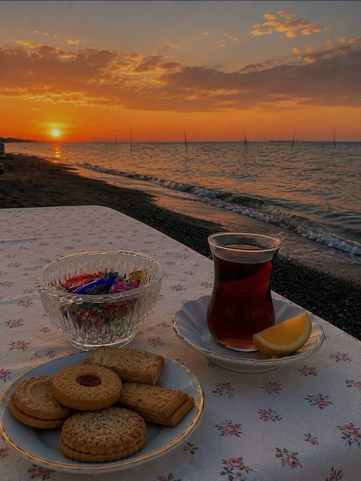 A serene sunset over the sea frames a traditional Turkish tea setup, featuring a glass of dark tea served with a lemon wedge on a saucer, a small bowl of candies, and a plate of assorted biscuits arranged on a floral tablecloth. The tranquil seaside scene contrasts with the post's promotion of Tria's decentralized yield generation, evoking a moment of relaxation that parallels the financial ease the platform promises.