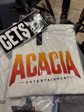 A white t-shirt or garment in plastic packaging displays the 'ACACIA ENTERTAINMENT' logo in orange-to-red gradient text, with a separate 'GETS' sticker or label featuring a skull-like face partially visible above it.