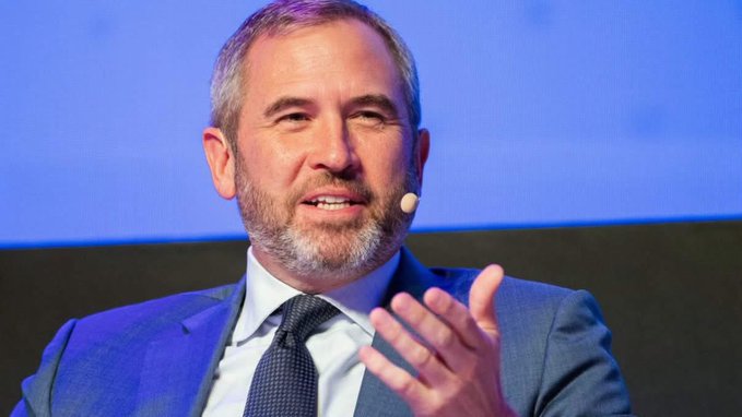 A middle-aged man with a beard in a blue suit and tie gestures with his hand while speaking, wearing a small earpiece microphone against a blue background.