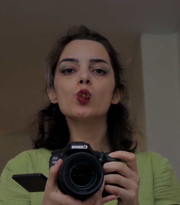 A woman with dark hair holds a Canon EOS camera up to her face while puckering her lips with a small yellow object inside, wearing a green top.