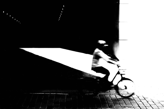 A high-contrast black and white image showing a motion-blurred figure riding a scooter along a brick pathway, with bright light illuminating a section of the background.