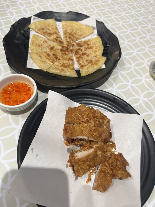 A meal on a patterned table featuring a sliced flatbread pancake on a black plate, a small dish of orange chili sauce, and crispy fried chicken pieces on parchment paper atop another black plate.