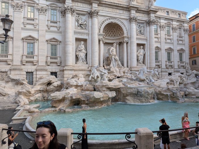 A view of the Trevi Fountain in Rome with its ornate baroque sculptures, turquoise water, and tourists gathered around.