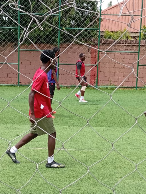 Three men in athletic wear play soccer on an artificial turf field, viewed through a chain-link fence.