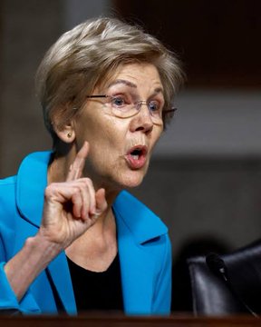 A close-up of an older woman with short blonde hair and glasses, wearing a teal blazer, gesturing with her index finger raised while speaking.