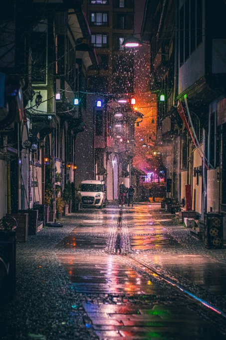 A rainy, neon-lit urban alleyway at night with colorful reflections on the wet pavement, featuring string lights overhead and a white car parked along the street.