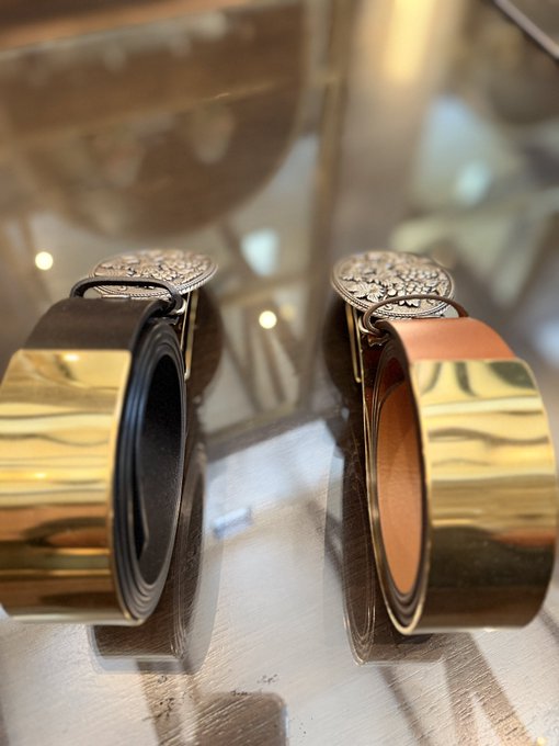 A close-up photograph of two luxury leather belts with large, ornate silver buckles coiled on a reflective glass surface.