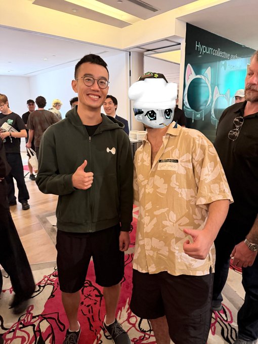 A smiling man wearing a "Hyperliquid" name tag and a cat-themed mask poses next to another man giving a thumbs up at what appears to be a Hyperliquid event in Singapore, with a "HypurrCollective" banner visible in the background. The caption ties into the post about meeting the Hyperliquid team and community, with the playful "Singapurr" reference matching the cat mask worn by the person on the right.