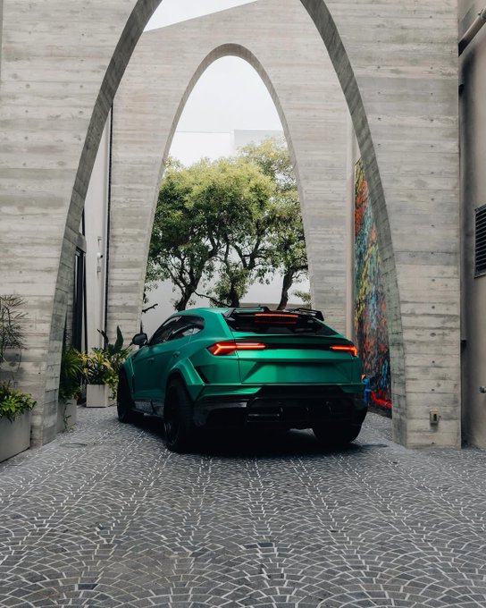 A matte green Lamborghini Urus SUV parked under a modern concrete archway on a cobblestone driveway, with a colorful mural and trees visible in the background.