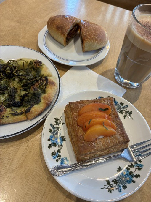 A wooden table displays a spread of food and drink: a slice of pizza topped with greens, a bread roll filled with chocolate, a tall glass of latte, and a layered pastry garnished with peach slices on a floral-patterned plate.