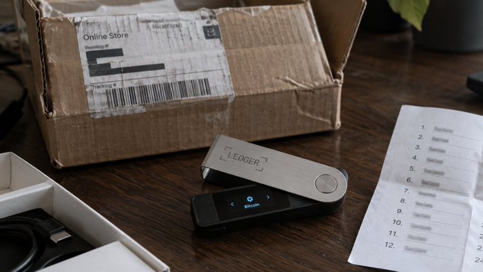 A Ledger hardware wallet being set up with Bitcoin on its screen, shown unboxed next to a shipping package and a recovery sheet on a wooden desk.