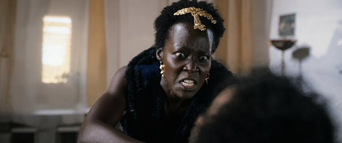 A dramatic scene of a woman with a gold headpiece showing an intense, angered facial expression while looking downward.