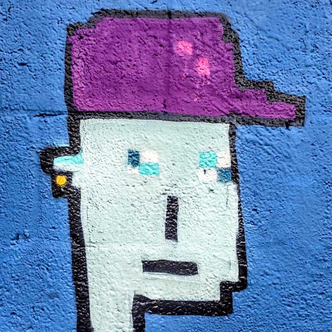 A street art mural on a blue wall depicting a pixelated white character with teal eyes wearing a purple cap.