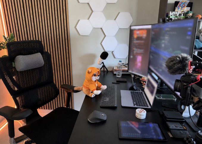 A modern home office and streaming setup featuring dual monitors, a laptop, keyboard, and a professional microphone with a furry windscreen, accompanied by a Shiba Inu plush toy in an orange hoodie—visually nodding to the $DOG memecoin referenced in the post about trading meme tokens.
