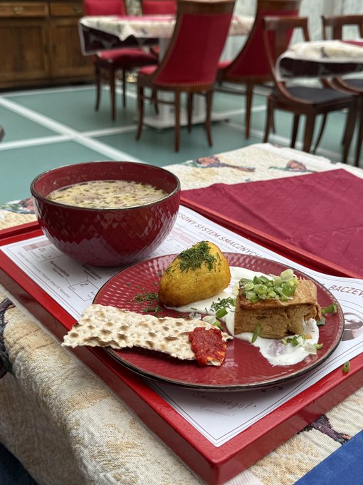 A bowl of creamy soup and a plate with what appears to be meat, potato, and matzah served on a red tray in a restaurant setting with red upholstered chairs.