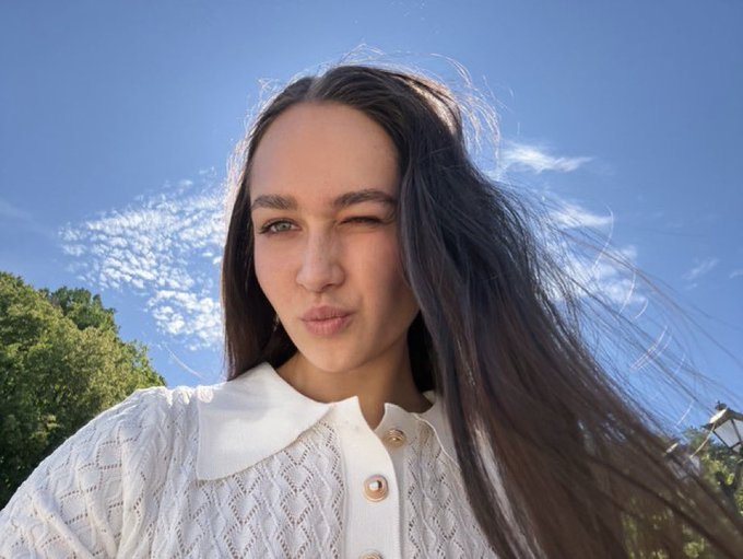 A selfie of a woman with long dark hair winking and making a kiss face outdoors against a blue sky.