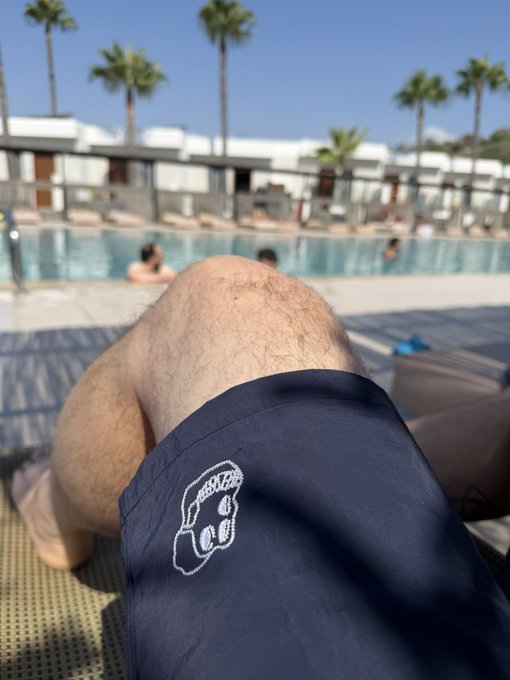 A first-person view of a person relaxing by a pool, showing their knee and navy blue shorts with a white embroidered logo, while other swimmers are visible in the background under palm trees.