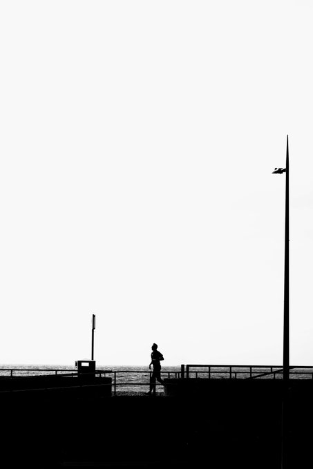 A minimalist black-and-white silhouette photograph of a person walking along a seaside promenade with a tall lamppost against a pale sky.
