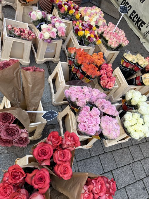 A vibrant flower stand displays colorful bouquets of roses and ranunculus with visible price tags (9€, 19€, 25€) and a sign reading "BLUMEN ECKE." The image is humorously used to symbolize the post's celebration of pumping the token "$swappy" from a $70k entry to a $1M market cap, jokingly comparing the flurry of financial activity to an abundant market blooming with profits.