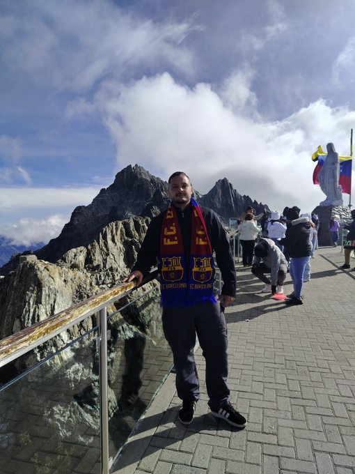 A man wearing a red and blue FC Barcelona scarf stands at a mountain overlook with a statue and flags in the background.